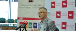 Mężczyzna w okularach siedzi przy stole z mikrofonami. W tle logo Politechniki Świętokrzyskiej.