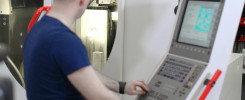 Mężczyzna w niebieskiej koszulce obsługuje panel sterowania maszyny CNC. Widoczny panel sterowania, ekran i część maszyny.