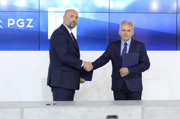 Dwóch mężczyzn w garniturach ściska sobie dłonie. Trzymają teczki. W tle logo PGZ.
