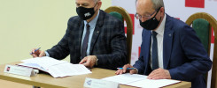 Dwóch mężczyzn w garniturach i maskach siedzi przy stole, podpisując dokumenty. W tle logo Politechniki Świętokrzyskiej.