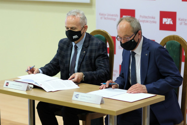 Dwóch mężczyzn w garniturach i maskach siedzi przy stole, podpisując dokumenty. W tle logo Politechniki Świętokrzyskiej.