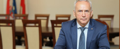 Mężczyzna w garniturze siedzi przy stole. W tle flagi i obrazy.