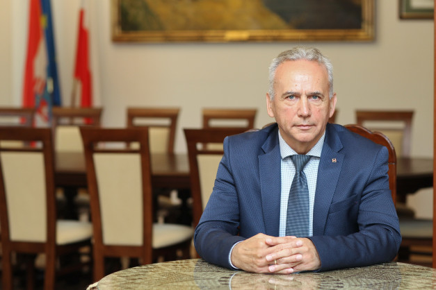 Mężczyzna w garniturze siedzi przy stole. W tle flagi i obrazy.