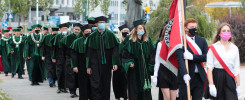 Grupa osób w togach akademickich i biretach maszeruje z flagą. W tle budynki i pomnik.
