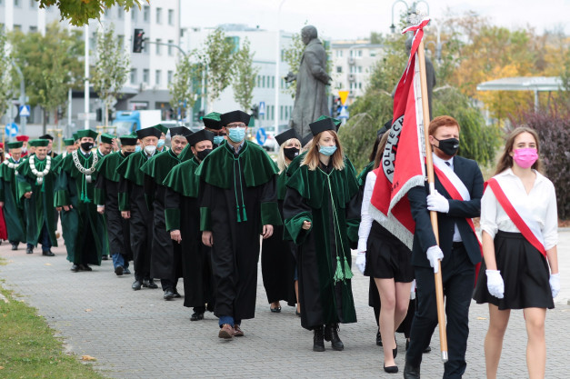 Grupa osób w togach akademickich i biretach maszeruje z flagą. W tle budynki i pomnik.