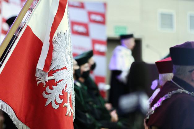 Flaga Polski z orłem, ludzie w togach i biretach. Kontekst: uroczystość akademicka lub państwowa.