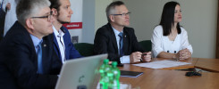 Cztery osoby siedzą przy stole. Na stole laptop, dokumenty, butelki z wodą. Kontekst: spotkanie.
