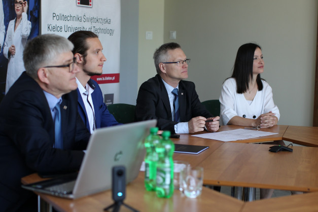 Cztery osoby siedzą przy stole. Na stole laptop, dokumenty, butelki z wodą. Kontekst: spotkanie.