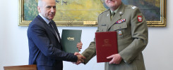 Dwóch mężczyzn, jeden w garniturze, drugi w mundurze wojskowym, wymienia uścisk dłoni. Trzymają dokumenty.