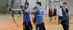 Łucznicy w hali sportowej, z łukami i strzałami. W tle boisko do koszykówki i tablica wyników.