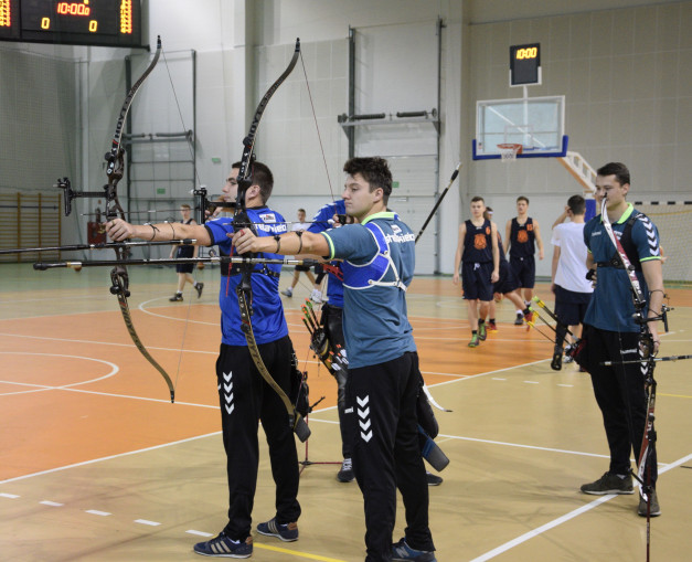Łucznicy w hali sportowej, z łukami i strzałami. W tle boisko do koszykówki i tablica wyników.
