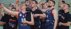 Drużyna koszykarzy świętuje zwycięstwo. Kluczowe obiekty: zawodnicy w strojach sportowych, ręce w geście radości, hala