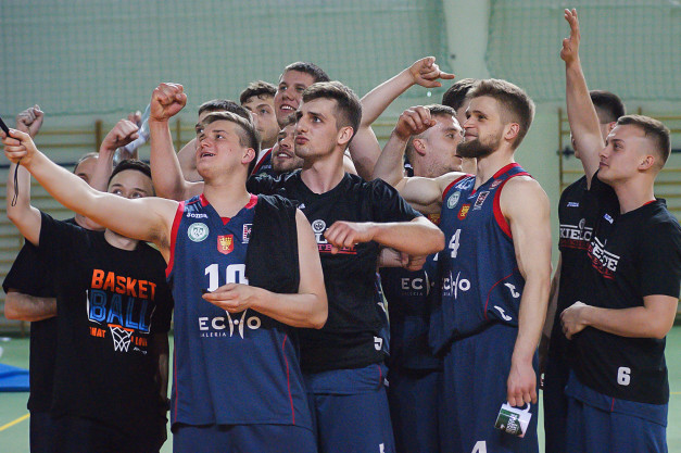 Drużyna koszykarzy świętuje zwycięstwo. Kluczowe obiekty: zawodnicy w strojach sportowych, ręce w geście radości, hala