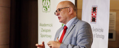 Mężczyzna w garniturze przemawia na tle banerów. Kluczowe obiekty: mężczyzna, okulary, garnitur, banery.