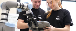 Dwóch ludzi w czarnych koszulkach z logo PSK patrzy na tablet. Obok nich znajduje się robot.