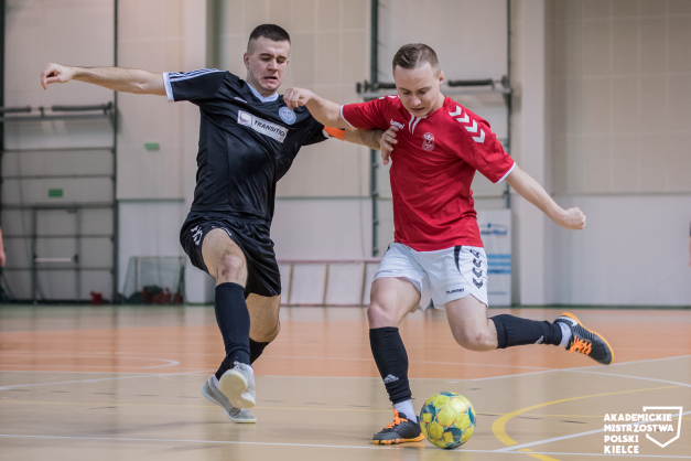 Dwóch piłkarzy walczy o piłkę na hali sportowej. Jeden w czarnej koszulce, drugi w czerwonej.