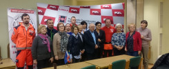 Grupa osób pozuje do zdjęcia na tle banerów. Widać ratownika w pomarańczowym stroju.