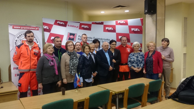 Grupa osób pozuje do zdjęcia na tle banerów. Widać ratownika w pomarańczowym stroju.