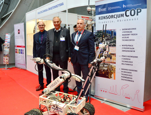 Trzech mężczyzn stoi obok robota i banerów. Kluczowe obiekty: robot, banery, mężczyźni. Kontekst: targi lub wystawa.