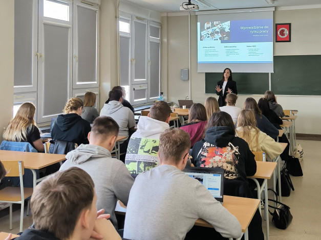 Nauczycielka prowadzi lekcję o rynku pracy. Uczniowie siedzą przy ławkach, niektórzy korzystają z laptopów.