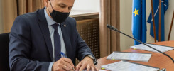 Mężczyzna w garniturze i masce podpisuje dokumenty. Na stole leżą dokumenty, mikrofon, a w tle flagi UE i NATO.