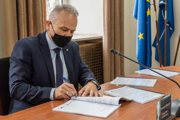 Mężczyzna w garniturze i masce podpisuje dokumenty. Na stole leżą dokumenty, mikrofon, a w tle flagi UE i NATO.