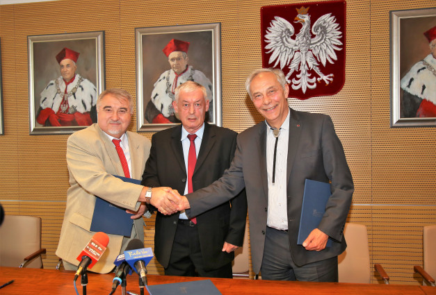 Trzech mężczyzn w garniturach ściska sobie dłonie. W tle portrety i godło Polski. Kontekst: uroczystość, spotkanie.
