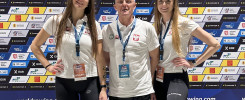 Trzy osoby w białych koszulkach z polskimi symbolami na tle baneru "2024 World Rowing Indoor Championships".