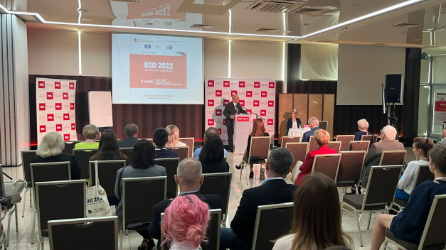 Konferencja BSD 2022. Mężczyzna przemawia na podium, widownia siedzi. Kluczowe obiekty: podium, ekran, widownia.