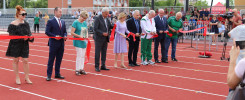 Osoby przecinają czerwoną wstęgę na bieżni stadionu. Kluczowe obiekty: ludzie, wstęga, bieżnia.