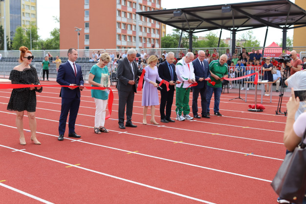 Osoby przecinają czerwoną wstęgę na bieżni stadionu. Kluczowe obiekty: ludzie, wstęga, bieżnia.