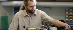 Mężczyzna z brodą i okularami pracuje nad dronem. Na stole znajdują się części elektroniczne i narzędzia.