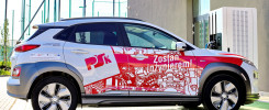 Biały samochód elektryczny z czerwonymi grafikami i napisem "Zostań inżynierem!". Obok ładowarka.