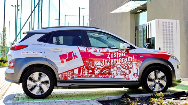 Biały samochód elektryczny z czerwonymi grafikami i napisem "Zostań inżynierem!". Obok ładowarka.
