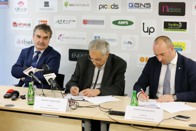 Trzech mężczyzn przy stole, jeden podpisuje dokument. W tle logo firm.