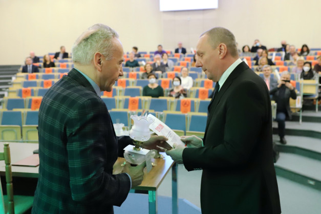 Dwóch mężczyzn w garniturach odbiera dyplom i lampę w sali wykładowej. W tle siedzą ludzie.