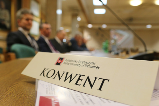 Tabliczka z napisem "KONWENT" i logo Politechniki Świętokrzyskiej. W tle siedzą mężczyźni w garniturach.