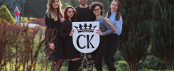 Grupa osób pozuje z okrągłym logo z koroną i literami "CK". W tle drzewa i trawnik.