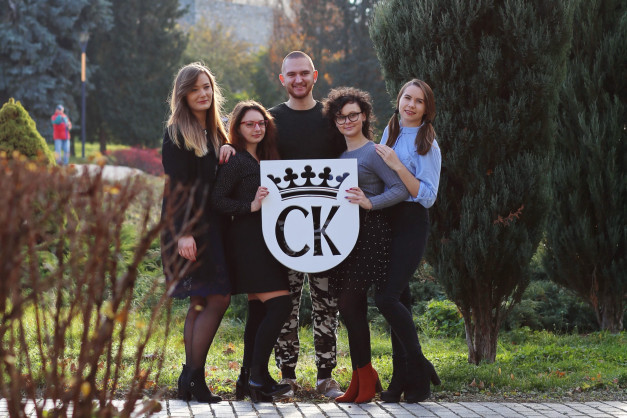Grupa osób pozuje z okrągłym logo z koroną i literami "CK". W tle drzewa i trawnik.