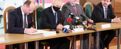 Czterech mężczyzn w garniturach siedzi przy stole, podpisując dokumenty.