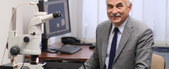Mężczyzna w garniturze siedzi przy biurku z mikroskopem, monitorem i telefonem. Kontekst: laboratorium.