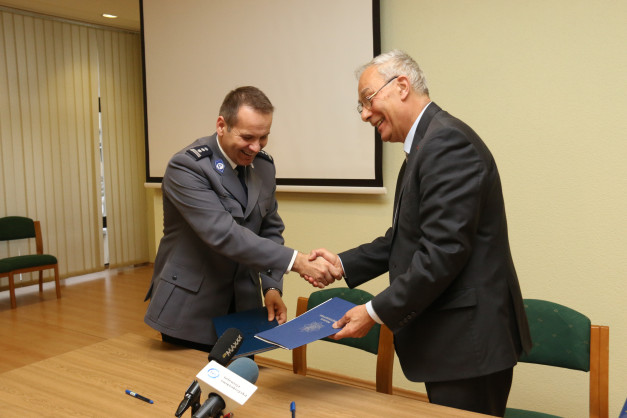 Dwóch mężczyzn, jeden w mundurze policyjnym, drugi w garniturze, ściska sobie dłonie. Na stole leżą dokumenty i mikrofony.