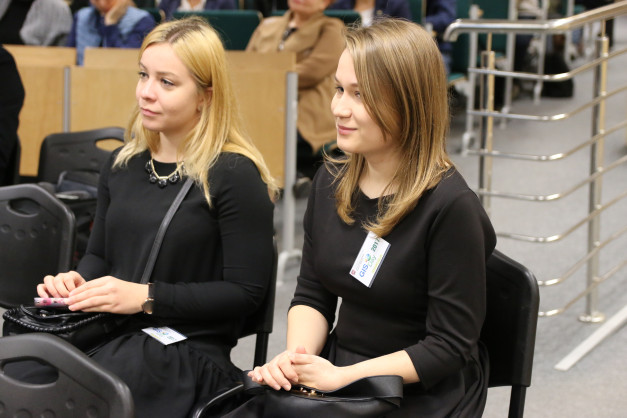 Dwie kobiety w czarnych strojach siedzą na krzesłach. Jedna z nich ma przypiętą plakietkę GIS Day 2017.