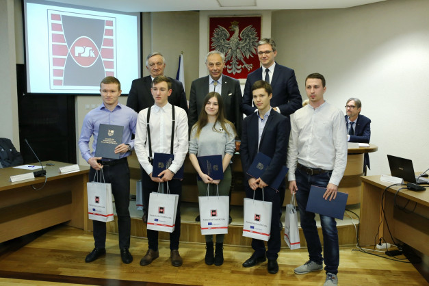 Osoby odbierają dyplomy i torby z logo Politechniki Świętokrzyskiej. W tle herb Polski i logo uczelni.