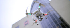Biały dron z zielonymi i czerwonymi światłami leci przed nowoczesnym budynkiem. Kluczowe obiekty: dron, budynek.