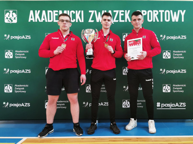 Trzech mężczyzn w czerwonych bluzach i czarnych spodniach pozuje z pucharem i dyplomem. Kontekst: Akademicki Związek Sportowy.