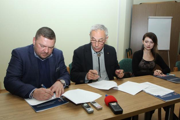 Trzech ludzi przy stole podpisuje dokumenty. Widać telefony, dyktafony i dokumenty.