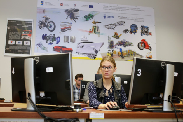Młoda kobieta pracuje przy komputerze. Na ścianie wiszą plakaty z grafikami pojazdów i maszyn.