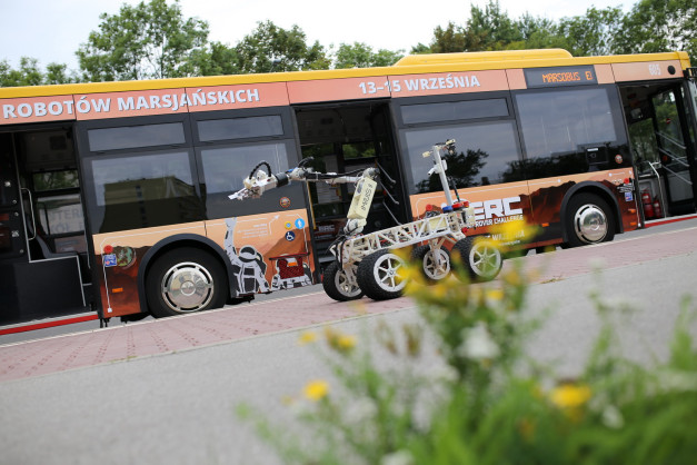 Autobus z napisem "ROBOTÓW MARSJAŃSKICH" i łazik marsjański. Kluczowe obiekty: autobus, łazik marsjański, robotyczne ramię.