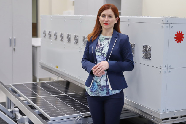 Kobieta w garniturze stoi obok paneli słonecznych i białej obudowy z wentylatorami. Kontekst: technologia, energia odnawialna.